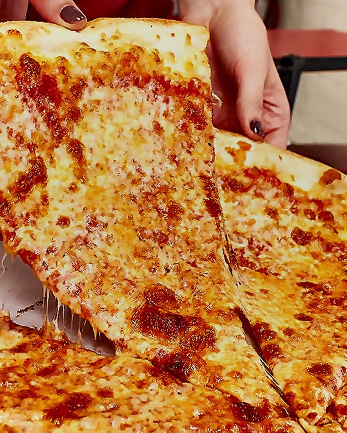 Hand lifting a big New York cheese slice from a whole pie
