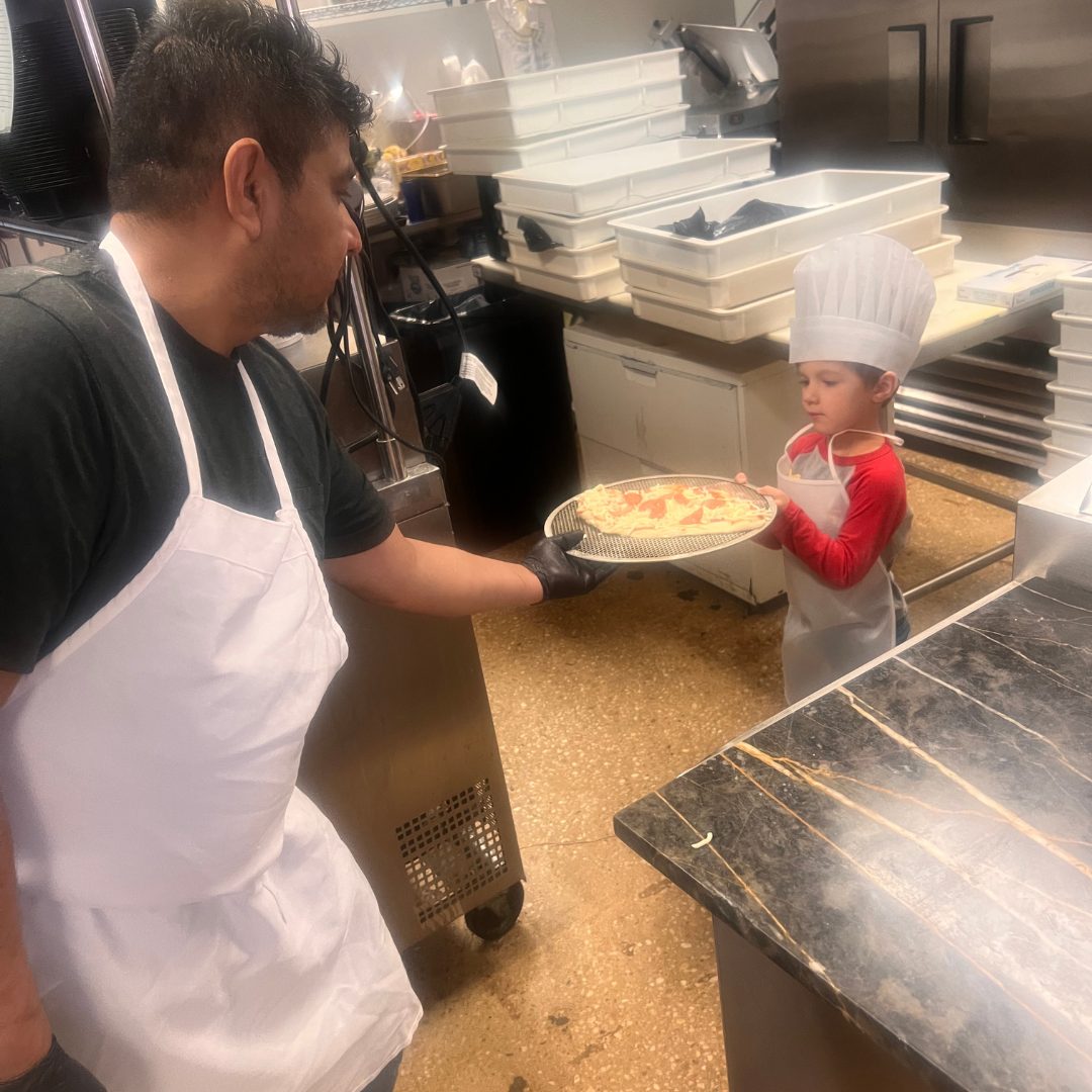 Gigi's chef handing a freshly topped pizza to a young party guest in a chef hat