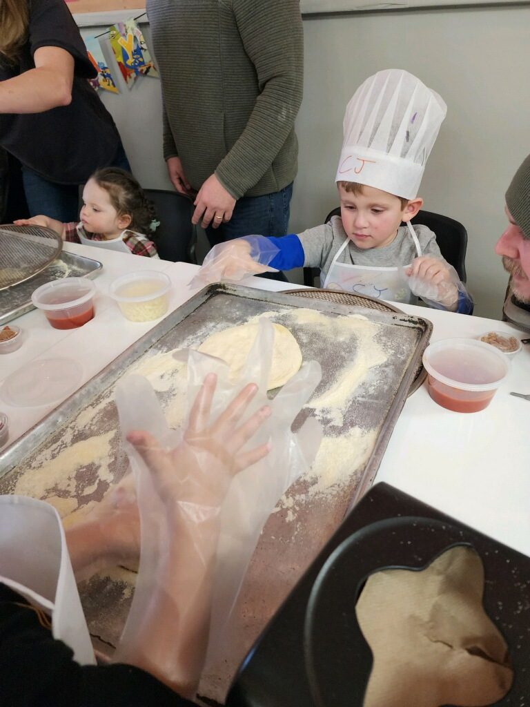 Children at the party table stretching pizza dough, with cups of sauce and cheese in front of them